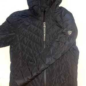 Svart dunjacka från Emporio Armani EA7 - Svart dunjacka från Emporio Armani EA7 med huva och snyggt mönstrad quiltning. Jackan har dragkedja framtill med logga och en patch på ärmen. Perfekt för kalla dagar och ger en clean, modern look. Passar XS/S