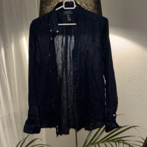 Blå linneskjorta från Polo Ralph Lauren - Snygg mörkblå skjorta i linne från Polo Ralph Lauren, storlek XL ca 175. Skjortan har klassisk krage, långa ärmar med manschett och knappar framtill. På bröstet finns den ikoniska röda Polo-loggan broderad. Perfekt för dig som gillar stilrena och bekväma plagg.