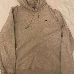 Beige hoodie med liten broderad logga - Säljer en beige hoodie med huva och dragsko. Tröjan har en liten broderad logga på bröstet och är tillverkad i mjukt material. Perfekt för en avslappnad stil.
