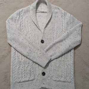 Ljusgrå stickad cardigan med knappar - Säljer en mysig ljusgrå stickad kofta med kabelstickat mönster och svarta prickar. Koftan har två stora knappar framtill, ribbade muddar och en bred krage. Perfekt för lager-på-lager och chill dagar. Storleken passar för S och M