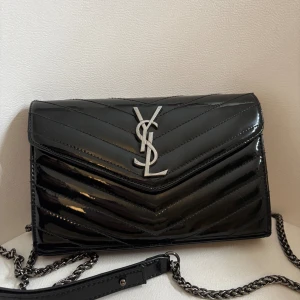 Svart YSL axelväska i lackskinn - Snygg svart axelväska från Saint Laurent i glansigt lackskinn med silverfärgad YSL-logga framtill. Väskan har kedjerem, quiltad design och magnetlås. Insidan har fack med dragkedja och plats för kort. Perfekt för dig som vill ha en lyxig och trendig accessoar.