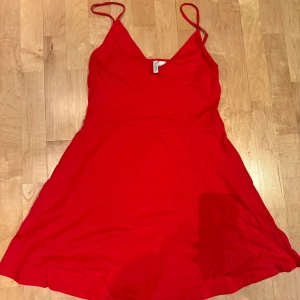 Röd kort klänning från H&M Divided - Enkel och snygg röd klänning från H&M Divided med smala axelband och v-ringning. Modellen är kort och har en lätt utställd kjol. Tillverkad i mjuk bomull, perfekt för varma dagar eller när du vill sticka ut lite extra.