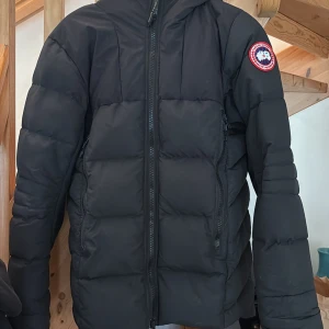 Svart dunjacka från Canada Goose  - Svart Canada Goose jacka med luva. Jackan har dragkedja fram och en logotyp på vänster ärm. Den har även reflexer på luvan och remmar på insidan. Om de behövs fler bilder så har jag det, kunde bara inte lägga upp alla här:)