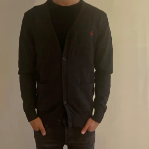 Mörkgrå kofta från Polo Ralph Lauren - Snygg mörkgrå kofta från Polo Ralph Lauren med klassisk röd logobrodyr på bröstet. Koftan har v-ringad hals, två framfickor och knäppning med svarta knappar. Materialet är mjukt och stickat, perfekt för lager på lager under höst och vinter.