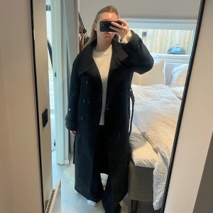 Svart lång populär kappa från Gina Tricot - Säljer en svart lång kappa från Gina Tricot i ullblandning. Modellen har bred krage, dubbelknäppning med stora svarta knappar och bälte i midjan. Kappan är helsvart, har lång ärm och en snygg slits bak för extra rörelsefrihet. (Modell - belted wool blend coat)