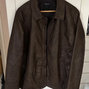 Brun skinnjacka L - Snygg brun skinnjacka från The Black Label Collection i storlek L. Jackan har klassisk krage, dragkedja framtill och två fickor med dragkedja. Materialet är strukturerat och ger en cool vintagekänsla. Perfekt för dig som vill ha en tidlös och stilren look.