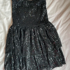 Svart glittrig paljettklänning som kan också användas som en topp - Supersnygg svart kortklänning med massor av glittrande paljetter över hela tyget. Klänningen har en volangkjol i flera lager som ger extra rörelse och festkänsla. Perfekt för dig som vill sticka ut med en glammig look. Materialet är mjukt och stretchigt syntet.