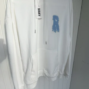 Vit hoodie från Risky med blå patch - Vit hoodie från Risky med stor huva och känguruficka. På bröstet sitter en blå patch i form av ett R med drip-effekt. Tröjan har långa ärmar och snörning vid halsen. Mjuk bomullskänsla och clean look, perfekt för dig som gillar streetwear. ( Helt ny, aldrig använt) väldigt sällsynt tröja 