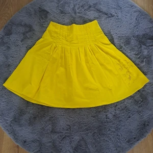 Gul kjol från The Dark Side of the Valley - Snygg gul kjol med hög midja och veckad design från The Dark Side of the Valley. Kjolen har en medium längd och ett subtilt broderat blommönster på ena sidan. Perfekt för dig som vill sticka ut med färg och stil.