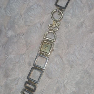 Klocka Mer info privat! - Säljer en unik klocka i guld med fyrkantig urtavla och strass runt både urtavlan och loggan. Armbandet har öppna fyrkantiga länkar och en cool design som sticker ut. Perfekt för dig som gillar bling och vill ha något som syns. Nya batterier behövs! Mer information privat!