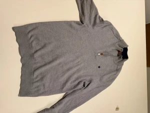 Grå merinoulltröja Morris half zip - Snygg grå långärmad tröja från Morris i 100% merinoull. Tröjan har en half zip-dragkedja vid halsen och ett diskret broderat emblem på bröstet. Ribbstickade muddar vid ärmslut och nederkant ger en clean look. Perfekt för kyliga dagar. Obs: tröjan har två små hål på baksidan som är lätta att sy ihop. Passar strl M