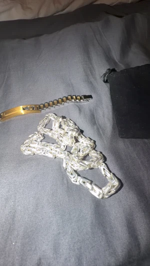 Smycken  - Bilderna visar en guldfärgad och silverfärgad länk, samt en svart tygpåse. Ingen väst syns på bilderna, så ingen beskrivning av väst kan ges.