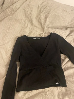 Svart omlottopp från Sister XXS - Säljer en svart omlottopp från Sister i storlek XXS. Toppen har ribbad struktur, lång ärm och djup v-ringning. Materialet är mjukt och stretchigt, perfekt för att skapa en snygg siluett. Passar dig som gillar stilrena och enkla plagg med lite edge.