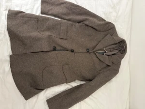 Brun kappa från Riley Shelby - Snygg brun kappa från Riley Shelby med klassisk krage och tre knappar framtill. Jackan har två stora fickor och är helfodrad med slits bak för extra rörelsefrihet. Perfekt för dig som gillar stilrena och tidlösa plagg. Trasigt innertyg vid vänster armhåla, inget som syns. Nypris 5000