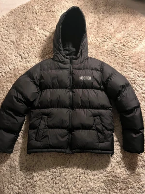 Svart pufferjacka från Hoodrich (XL barn storlek 13-15) - Svart pufferjacka från Hoodrich i barnstorlek XL  med huva och dragkedja. Jackan har broderad logga på bröstet och ryggen, samt två sidofickor. Perfekt för kalla dagar med sin tjocka och quiltade design.