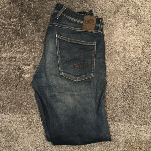 Replay mörkblå jeansbyxor slim fit - Snygga mörkblå jeansbyxor från Replay med klassisk femficksdesign och raka ben. Jeansen har diskreta slitningar, kontrastsömmar och läderpatch bak i midjan. Perfekt för dig som gillar en stilren och tidlös look. Ny Skick!