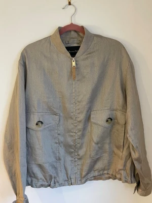  bomberjacka från Massimo Dutti - Snygg metallisk/guldig bomberjacka från Massimo Dutti med dragkedja framtill och stora knappar på fickorna och ärmsluten. Jackan har en stilren krage och är tillverkad i ett lätt linnematerial, perfekt för vår och sommar.