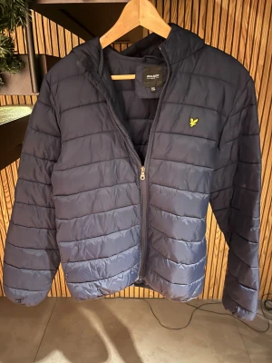 Mörkblå pufferjacka från Lyle & Scott - Snygg mörkblå pufferjacka från Lyle & Scott med huva och gul logga på bröstet. Jackan har horisontella quiltade paneler, dragkedja framtill och är tillverkad i polyester. Perfekt för kyliga dagar och enkel att matcha med din stil.