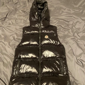 Svart dunväst från Moncler med huva - Säljer en svart, glansig dunväst från Moncler med huva och dragkedja framtill. Västen har två praktiska fickor med dragkedja och Moncler-logga på bröstet. Perfekt för att addera lite extra värme och stil till din outfit.
