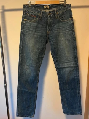Hilfiger Denim blå jeans straight fit - Blå jeans från Hilfiger Denim i klassisk straight fit med fem fickor och snygga slitningar. Jeansen har bruna sömmar, läderpatch med logga bak och detaljer vid fickorna. Perfekt passform och tidlös stil för dig som gillar denim. Strl. 32/34