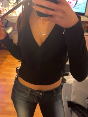 Svart omlottopp från ONLY - Säljer en svart omlottopp från ONLY i storlek M. Toppen har lång ärm, croppad passform och snygg v-ringning. Den är ribbad och har ett knyt i sidan som detalj. Perfekt att styla med jeans eller kjol för en cool look.