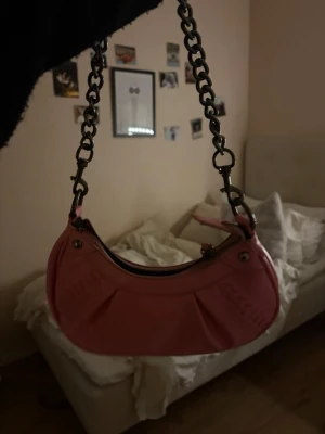 Rosa axelväska med kedja - Säljer en snygg rosa axelväska med grov metallkedja som axelrem. Väskan har en rundad form, dragkedja upptill och dekorativa veck på framsidan. Materialet är blankt syntet som ger en cool vibe. Perfekt för dig som gillar färg och vill sticka ut.