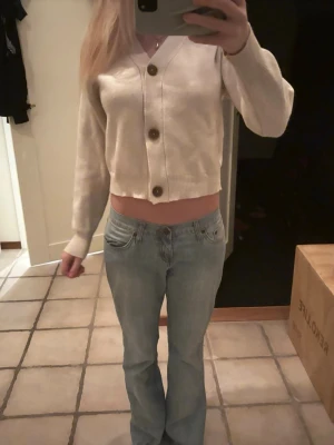 Beige croppad kofta med stora knappar - Snygg beige croppad kofta med v-ringning och tre stora bruna knappar framtill. Tröjan har lång ärm och är gjord i ett mjukt stickat material som känns skönt mot huden. Perfekt för dig som gillar en chill och trendig stil.