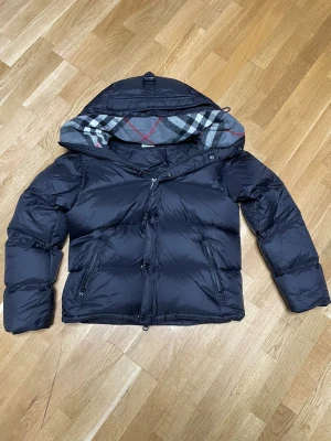 Burberry  - Svart pufferjacka från Burberry med avtagbara ärmar, vilket gör den till en väst. Jackan har klassiskt rutigt foder i huvan, dragkedja och knappar framtill samt två sidofickor med dragkedja. Materialet är glansig polyester och dunfyllning. Burberry-logga på ena ärmen.
