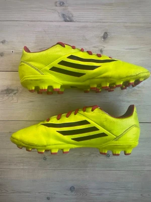 Gula Adidas fotbollsskor med röda detaljer - Säljer ett par riktigt snygga Adidas fotbollsskor i neon-gult med svarta ränder på sidorna och röda snören. Skorna har röda dobbar och insida, samt klassisk Adidas-logga under sulan. Tillverkade i syntetmaterial för bästa känsla på planen.