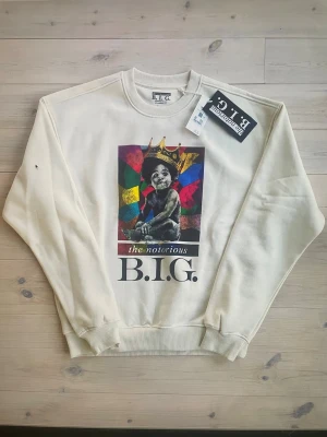 The Notorious B.I.G. Sweatshirt – Oversized Unisex - Riktigt fet sweatshirt för dig som älskar hiphop och legenden Biggie Smalls. Tröjan är helt ny med taggar kvar och kommer i en stilren off-white färg med ett ikoniskt tryck av The Notorious B.I.G. på framsidan. Oversized passform, rund halsringning och mjuk borstad insida gör den både grymt bekväm och sjukt snygg.  Kvalitén är hög och tyget är tjockt och följsamt – en tröja som verkligen håller. Officiell merch från Merch Traffic / Biggie Merch.  Storlek: Small Skick: Ny, aldrig använd