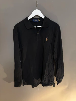 Svart långärmad piké från Polo Ralph Lauren - Svart långärmad pikétröja från Polo Ralph Lauren i modellen Custom Slim Fit. Klassisk krage, två knappar framtill och broderad logga på bröstet. Tillverkad i bomull och passar perfekt till en stilren och avslappnad look.