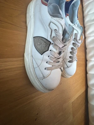 Philippe Model vita sneakers med blå detalj  - Säljer ett par vita Philippe Model sneakers med blå och röda detaljer vid hälen och silverfärgad logotyp på sidan. Skorna har snörning och är tillverkade i skinn med platt sula och rund tå. Perfekta för dig som gillar fransk streetstyle. Saknar sula på vänster sko