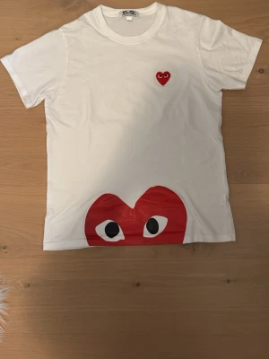 Vit Comme des Garçons Play t-shirt - Vit t-shirt från Comme des Garçons Play med det ikoniska röda hjärtat med ögon broderat på bröstet och ett stort tryckt hjärta längst ner. Klassisk rund hals och korta ärmar. Tillverkad i mjuk bomull.