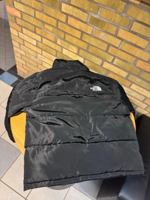 Svart pufferjacka från The North Face - Säljer en svart pufferjacka från The North Face med hög krage och två sidofickor med dragkedja. Jackan har en clean look med logga på bröstet och är tillverkad i glansig polyester. Perfekt för kalla dagar och riktigt snygg till streetwear.