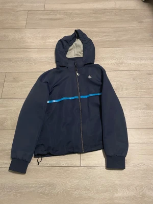 Blå vindjacka från Calvin Klein Jeans - Snygg marinblå vindjacka från Calvin Klein Jeans med huva och dragkedja. Jackan har en blå rand över bröstet och rygg, samt stort CK-logotryck på ryggen. Muddar vid ärmslut och justerbar nederkant. Perfekt för dig som gillar streetwear och vill ha något stilrent.