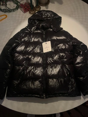 Svart Moncler Maya pufferjacka med huva - Svart pufferjacka från Moncler med glansig finish, huva och dragkedja framtill. Jackan har Moncler-logga på ärmen, två sidofickor med dragkedja och ett coolt serietidningsmärke på insidan. Perfekt för dig som vill ha en varm och snygg jacka med street vibe. Jackan är i storleken L men funkar lika bra som Medium om man tar en hoodie under. Jackan har svart insida och jackan är helt oanvänd. Taggen funkar. Skriv om ni har frågor/vill ha fler bilder.