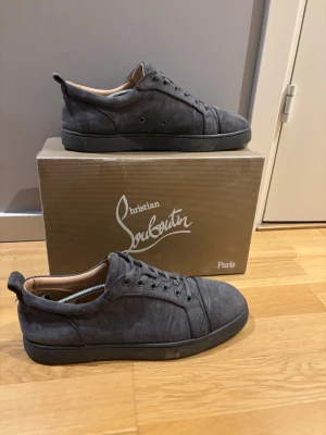 Grå sneakers från Christian Louboutin - Snygga grå sneakers från Christian Louboutin med klassisk röd sula och diskret logga. Skorna är tillverkade i mocka med rund tå och snörning. Insidan är beige och sulan är platt. Perfekt för dig som gillar exklusiva märken och stilren design.