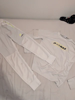 Vit Nike Air Max dress - Nike Air Max mjukisbyxor i vitt med gul neon-detalj längs benet och tryckt logga på låret. Byxorna har elastisk midja och är gjorda i mjukt bomullsmaterial, perfekt för chill eller träning. Snygg och sportig look med Air Max branding. Strl S