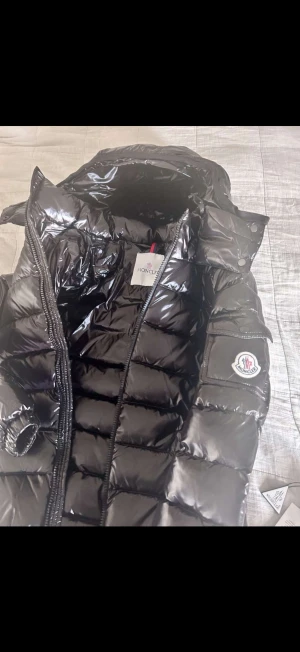 Svart Moncler pufferjacka med huva - Säljer en svart, glansig pufferjacka från Moncler med huva och dragkedja framtill. Jackan har quiltad design, Moncler-logga på ärmen och praktiska fickor. Perfekt för kalla dagar och har en riktigt snygg streetstil. Ingår (dustbag) aldrig använd. 