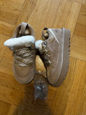 UGG LOWMEL - Snygga beige sneakers från UGG med grov platt sula och rund tå. Skorna har ett fluffigt vitt pälsfoder och mönstrade snören i beige och vitt. Ovandelen är i mocka och textil, perfekt för kalla dagar. Extra skosnören medföljer. SITTER OFAST 1-2 STORLEKAR MINDRE