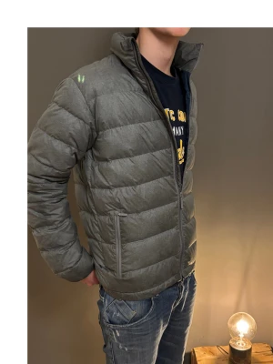 Grå pufferjacka med dragkedja - Säljer en stilren grå pufferjacka med hög krage och två sidofickor med dragkedja. Jackan har quiltad design och är perfekt för kyligare dagar. Passformen är normal och jackan är helfärgad utan mönster. Grym att slänga på sig när det är kallt ute!