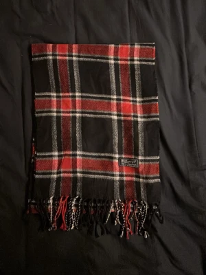 Rutig halsduk från Burberrys - Säljer en klassisk rutig halsduk från Burberrys i svart, rött och vitt. Halsduken är tillverkad i 100% lammull och har fransar längst ner. Perfekt för dig som gillar modern och klassisk stil och vill ha något som sticker ut lite extra. Inga defekter. Fraktar inom 24h. Hör av dig vid frågor eller funderingar🤩😃👍