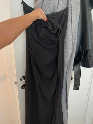 Nyårs kläning - Säljer en elegant svart långklänning med en stor dekorativ rosett framtill. Klänningen har en stilren och draperad design som ger en snygg siluett. Perfekt för dig som vill sticka ut med en unik detalj på festen.