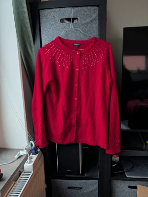 Röd kofta med pärlor från Exlindexlind - Snygg röd kofta från Exlindexlind i storlek 38/40. Koftan har rund hals, långa ärmar och knappar framtill. Dekorerad med pärlor i mönster över axlar och ärmslut som ger extra bling. Perfekt för dig som vill sticka ut med färg och detaljer.