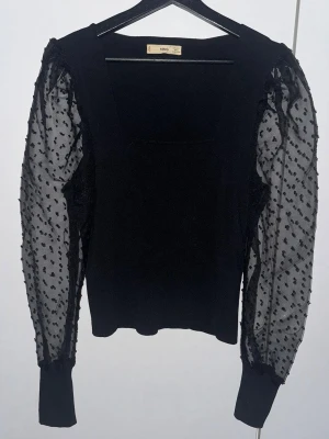 Svart topp med transparenta prickiga ärmar MNG - Säljer en svart topp från MNG med transparenta, långärmade meshärmar dekorerade med små sammetsprickar. Kroppen är i mjukt tyg och har en rak, något croppad passform. Ärmarna avslutas med breda muddar för en snygg kontrast.