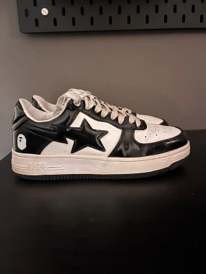 Svarta och vita BAPE STA sneakers - Säljer ett par BAPE STA sneakers i svart och vitt med klassisk stjärndesign på sidan. Skorna har snörning, perforerad tå och platt sula. Märkets logga syns tydligt på hälen och tungan. Materialet är blankt skinn och de har en rund tå. Perfekt för dig som gillar streetwear.