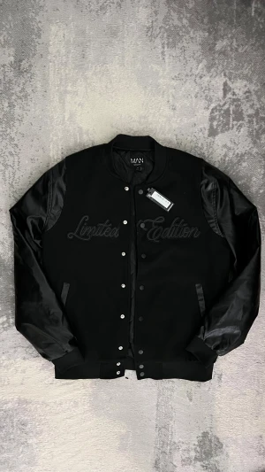 Svart bomberjacka från boohooMAN - Svart bomberjacka med broderad text 'Limited Edition' på bröstet. Jackan har blanka ärmar i syntetmaterial och quiltat foder på insidan. Stängs med knappar framtill och har ribbade muddar vid hals, ärmslut och nederkant. Perfekt för dig som vill ha en clean och stilren look.