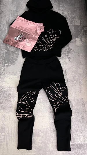 Syna world tracksuit  - Säljer min snygga och nästan oanvända Syna world limited edition tracksuit svart och rosé guld. Byxorna har dragkedja nere och man kan välja att ha öppet/flare eller stängt. Påse och accessoarer ingår.
