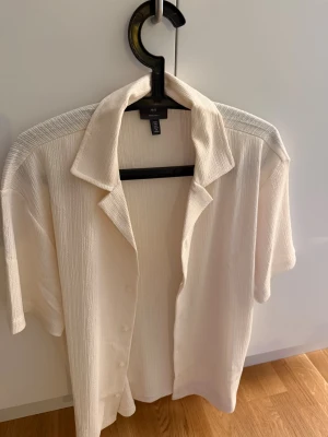 Vit kortärmad skjorta  - Säljer en vit skjorta från H&M med korta ärmar och krage. Skjortan har en snygg struktur i tyget och knappar framtill. Perfekt för dig som gillar en clean och chill stil. Passar dig som vill ha en enkel men ändå stilren look.