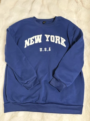 Blå sweatshirt med New York-tryck - Säljer en blå sweatshirt med vit text 'NEW YORK U.S.A' på bröstet. Tröjan har rund hals, långa ärmar och ribbade muddar vid ärmslut och nederkant. Perfekt för dig som gillar streetstyle och vill ha en enkel men snygg look.